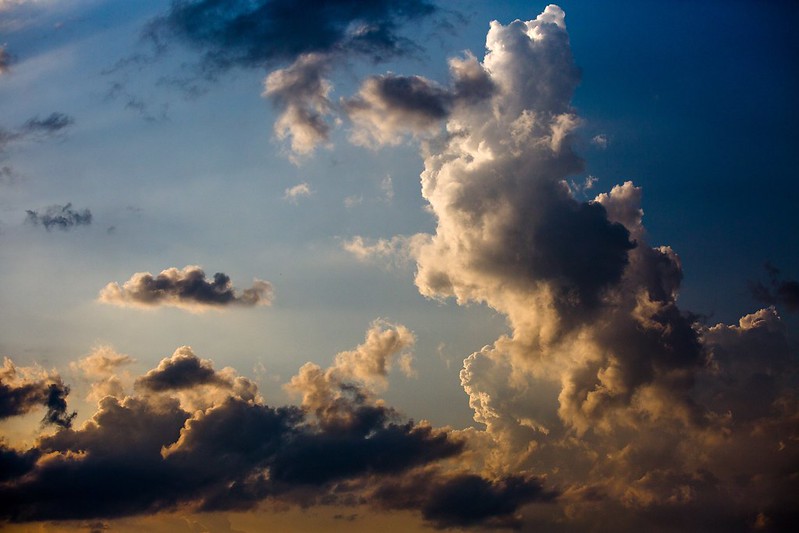 A photo of a towering cloud formation lit in white, blue, and rust by sunlight from the left with blue sky behind