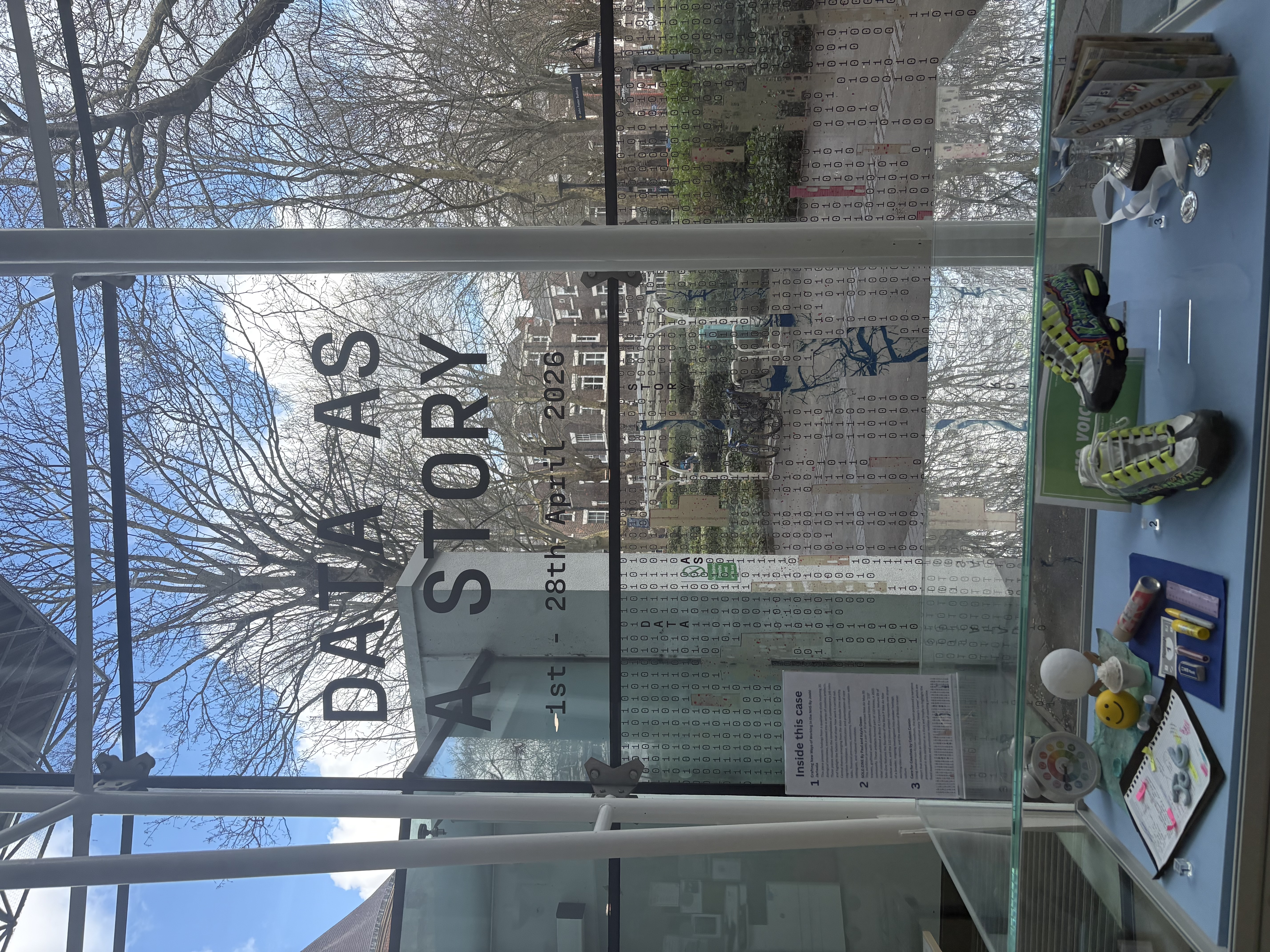 Display case from the 'Data as a Story' exhibition featuring personal objects and data-inspired artifacts, with the exhibition title visible on the glass wall behind
