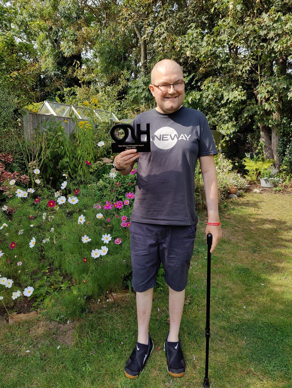Martin Paul Eve stands smiling broadly in front of wildflowers
                 and trees. He wears a Janeway T-shirt, holds the 2023 OLH
                 Award in one hand, and uses a cane for support with the other
                 hand.