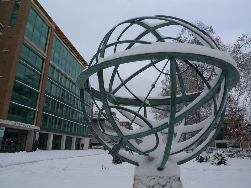 Photo of an outdoor sculpture of an abstract planet with
                concentric metal circles, covered in snow, with Birkbeck,
                University of London, in the background