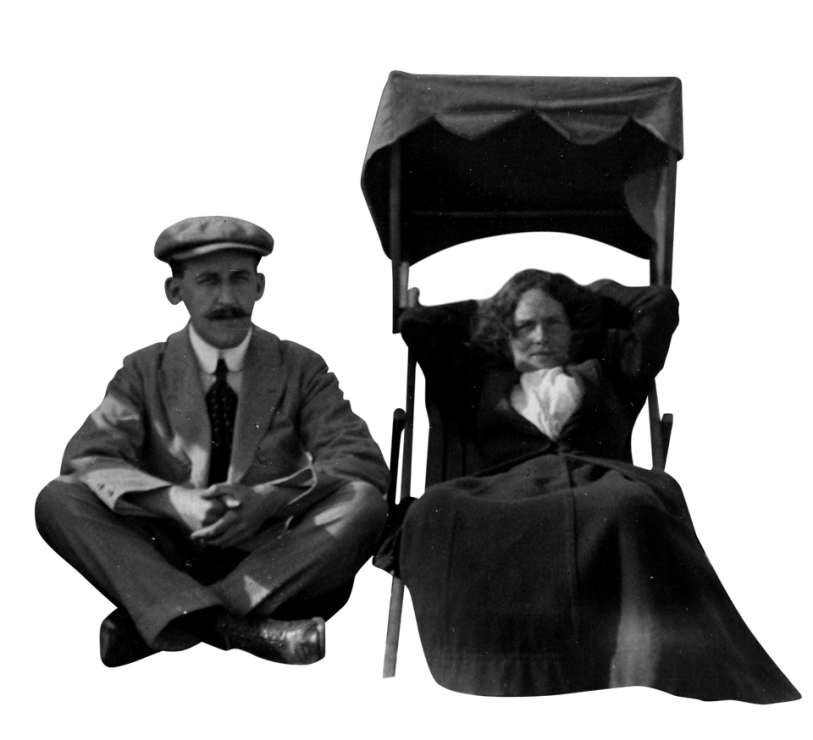 1930s photograph in which a grumpy woman slouches in a beach chair that has a high-back canopy to shield her from the sun, and a man in a suit with rounded collar sits cross-legged beside her